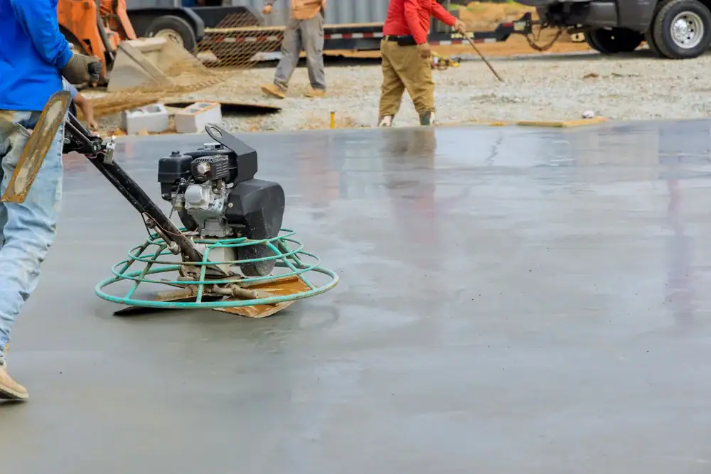 A construction worker uses a power trowel machine to smooth and finish a large, freshly poured concrete surface at an outdoor construction site. Other workers and vehicles are visible in the background.