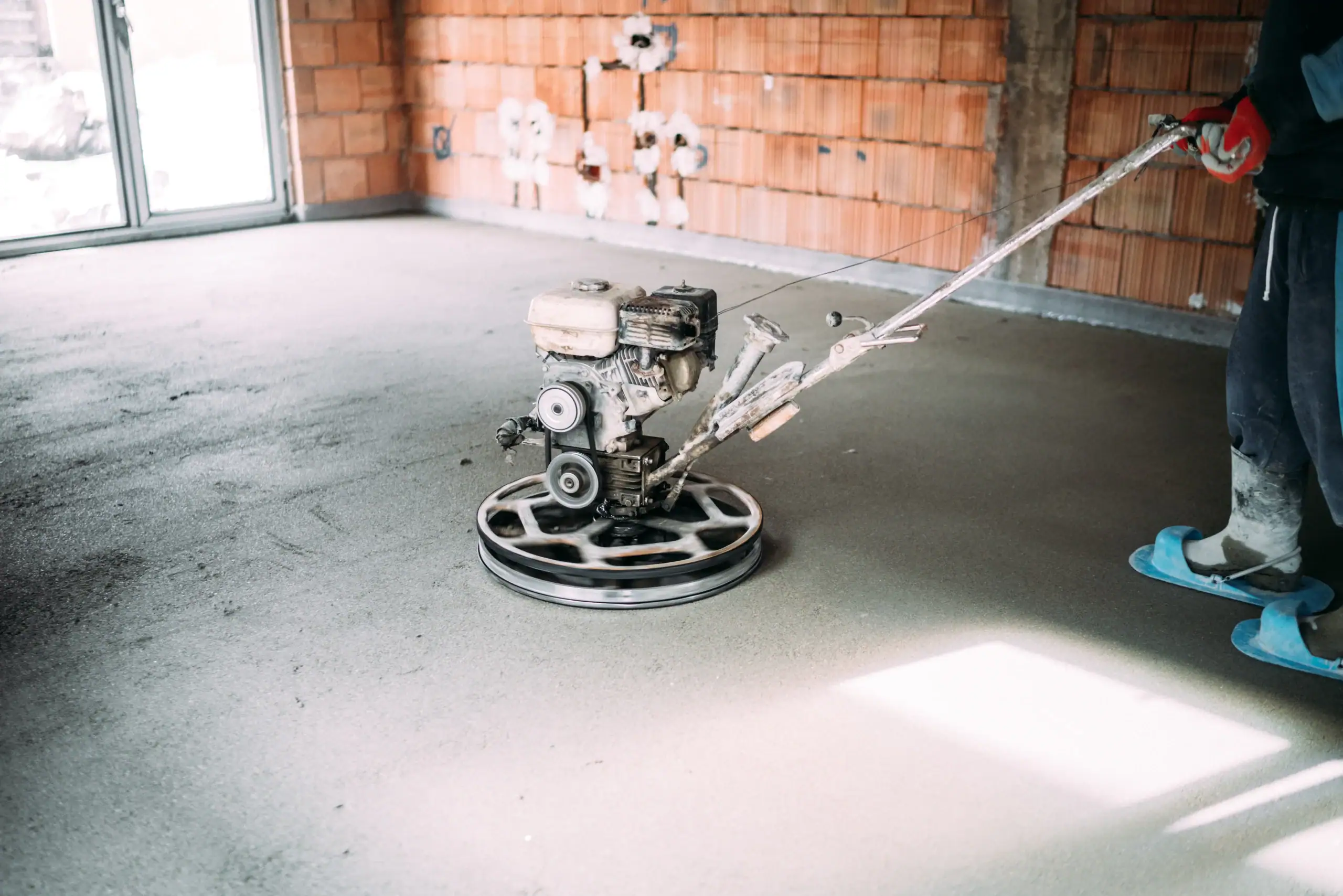 A construction worker uses a power trowel to smooth freshly poured concrete on the floor of a partially built room with exposed brick walls.