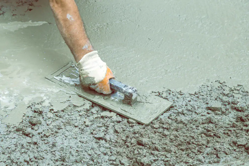 A close-up of a gloved hand smoothing wet concrete with a metal trowel, leveling the surface during construction work.