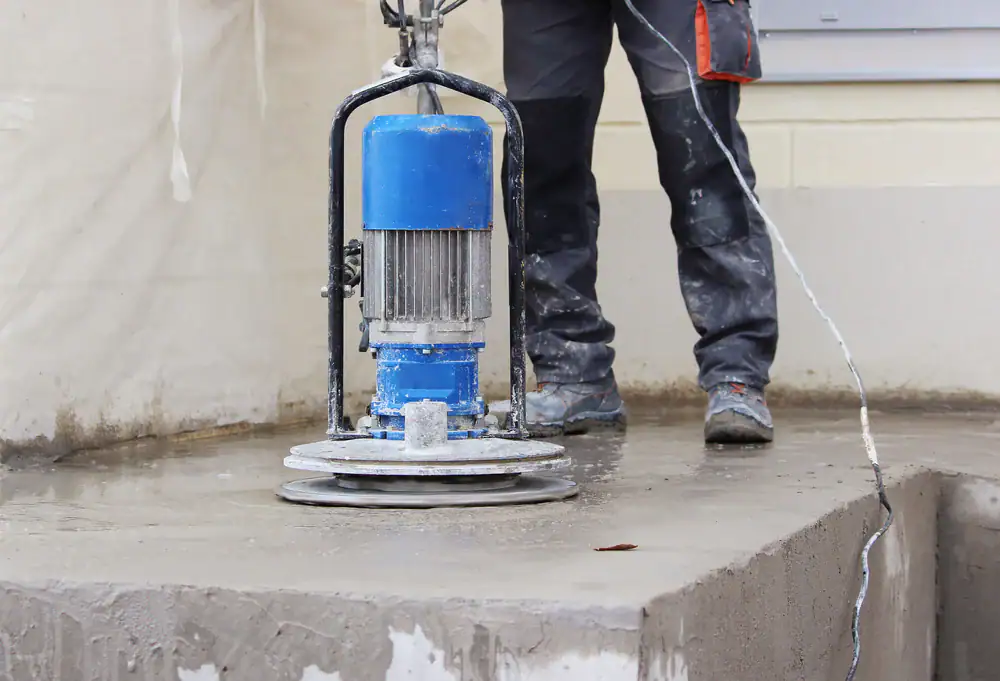 A worker uses a blue power trowel to smooth a concrete surface. The worker's lower body is visible, wearing work pants and boots, with the trowel spinning on a large, raised concrete slab.