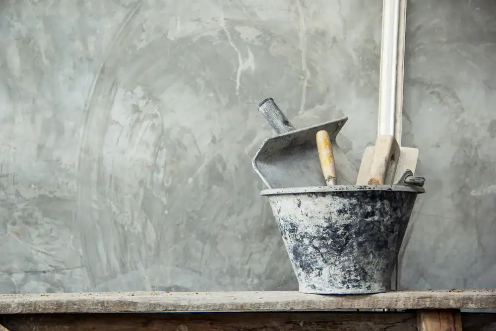 A metal bucket with plastering tools, including a trowel and spatulas, sits on a wooden surface against a gray, textured wall.