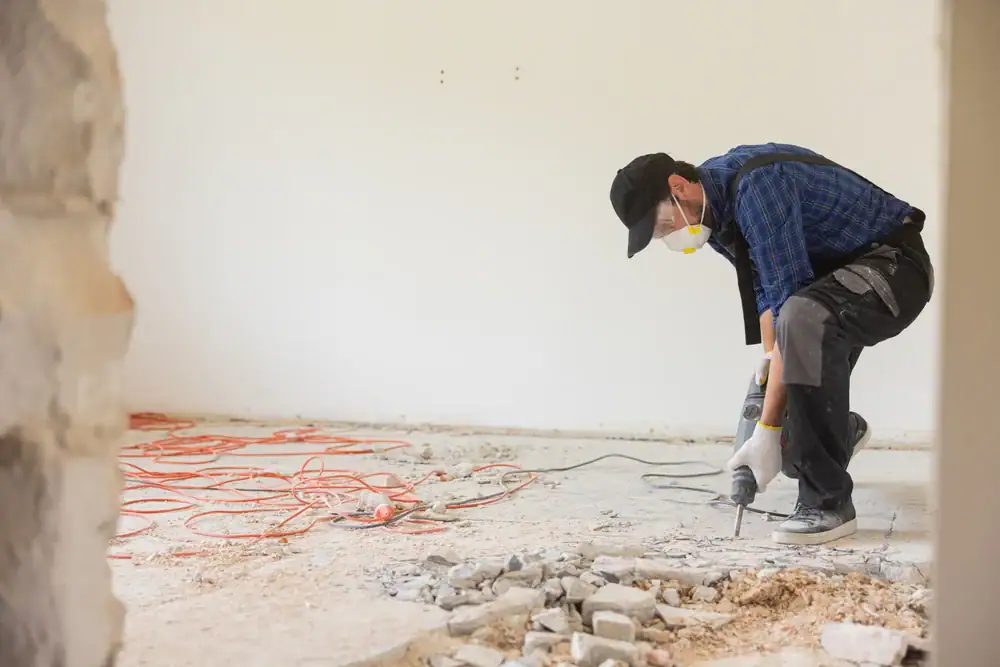 A construction worker wearing a mask and cap uses a power tool to break up concrete on a dusty floor in a room under renovation, with orange extension cords scattered in the background.