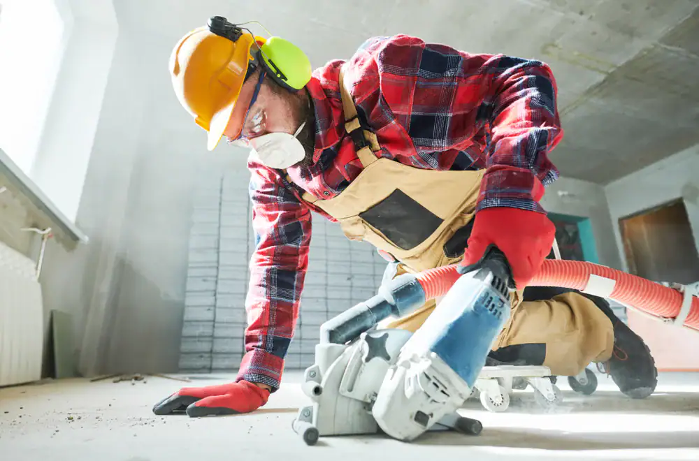 A construction worker in safety gear&mdash;hard hat, ear protection, goggles, mask, gloves, and overalls&mdash;operates a floor grinder on a dusty indoor site, kneeling on the ground while working.