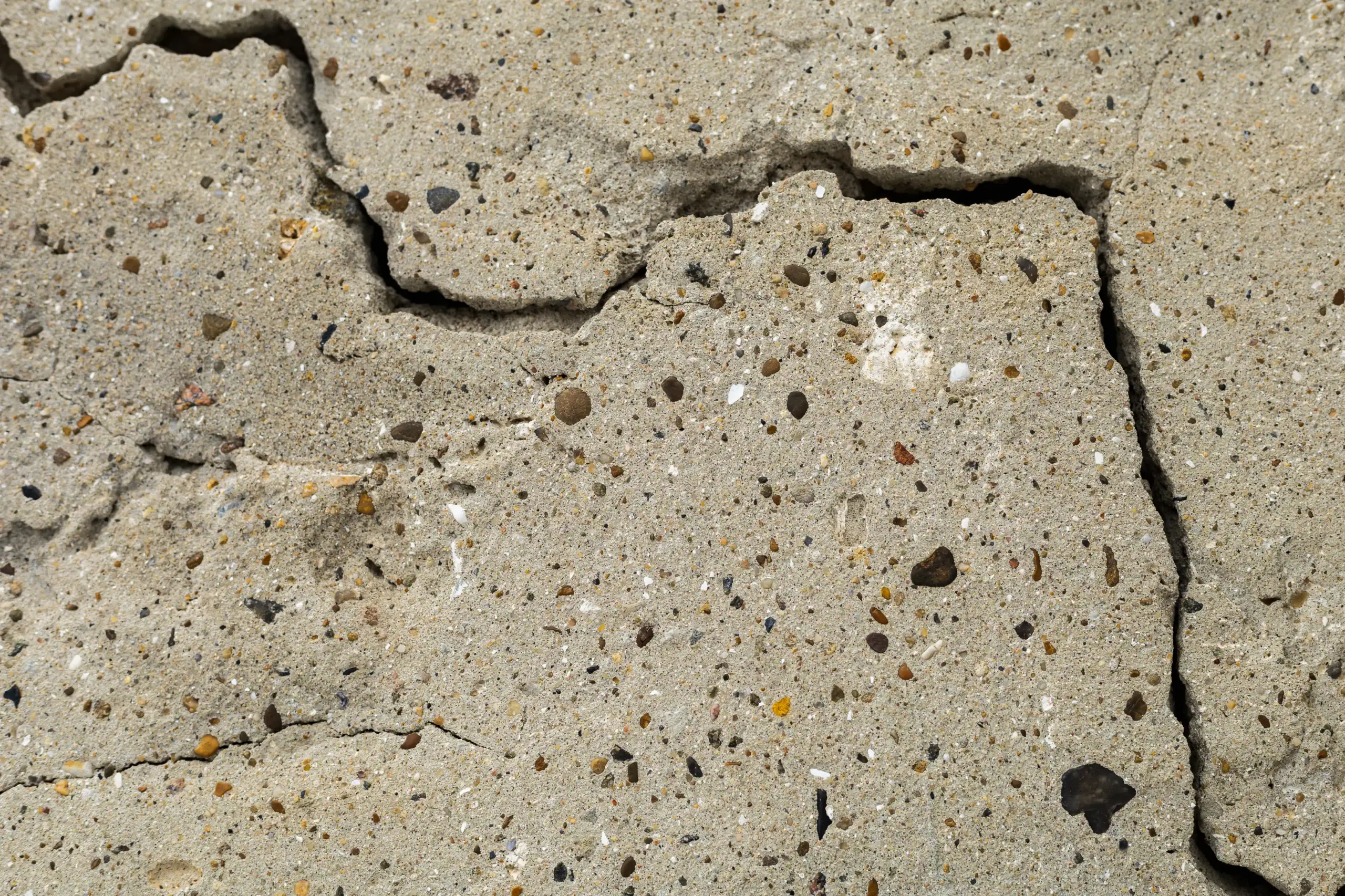 A close-up of a cracked concrete surface, showing rough texture, small stones, and sand embedded in the material. The crack runs irregularly across the image.