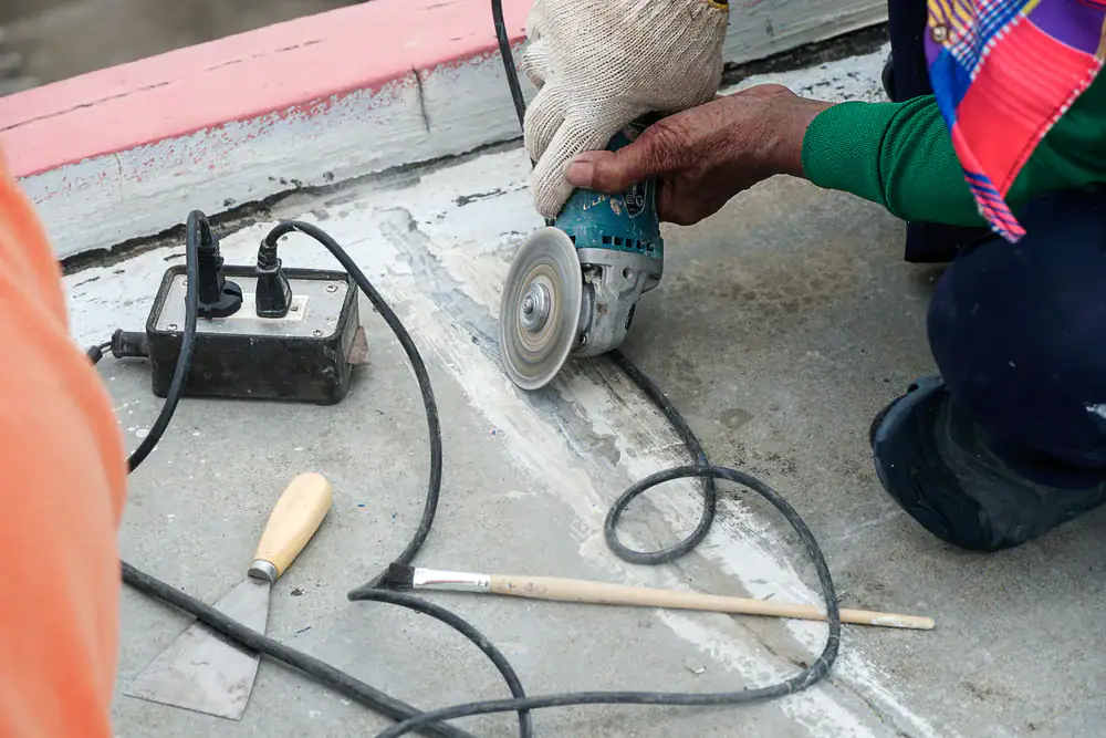 A person wearing gloves uses an angle grinder to cut a groove in a concrete surface. Nearby are a paintbrush, a chisel, and a power strip.