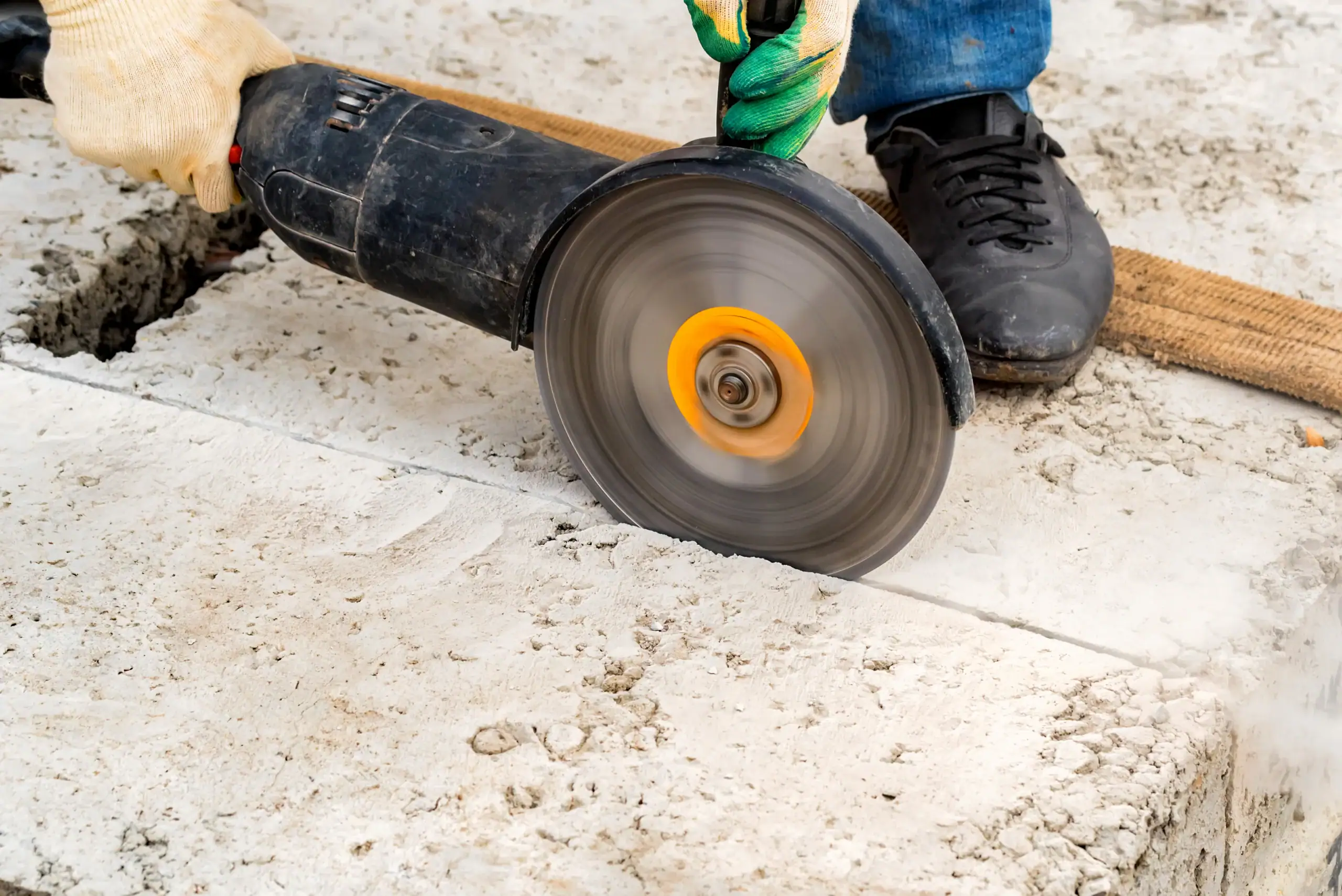 A person wearing gloves and jeans uses an angle grinder to cut through a concrete slab, with the spinning blade in motion and dust around the cutting area.