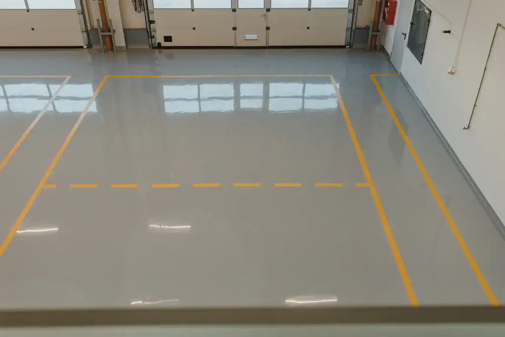 Empty industrial garage with a shiny grey floor, surrounded by yellow lines and a dashed yellow line in the center. Large windows reflect on the floor, and garage doors are visible in the background.