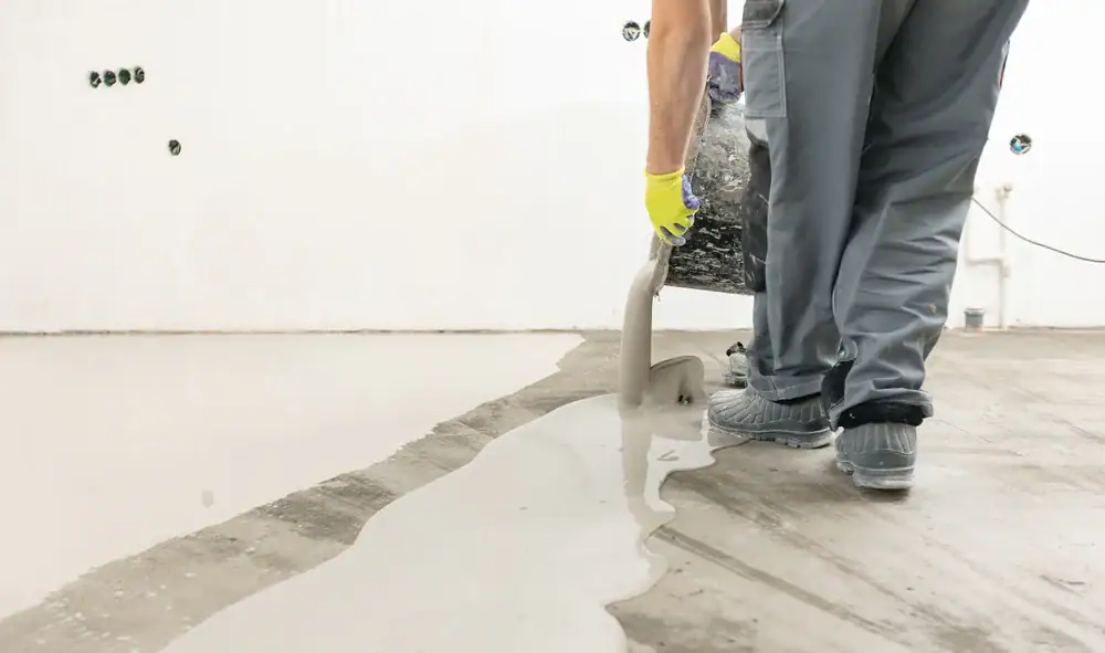 A person wearing gloves and work pants pours liquid concrete or self-leveling compound onto a bare floor, preparing the surface for finishing in a clean, white-walled room.