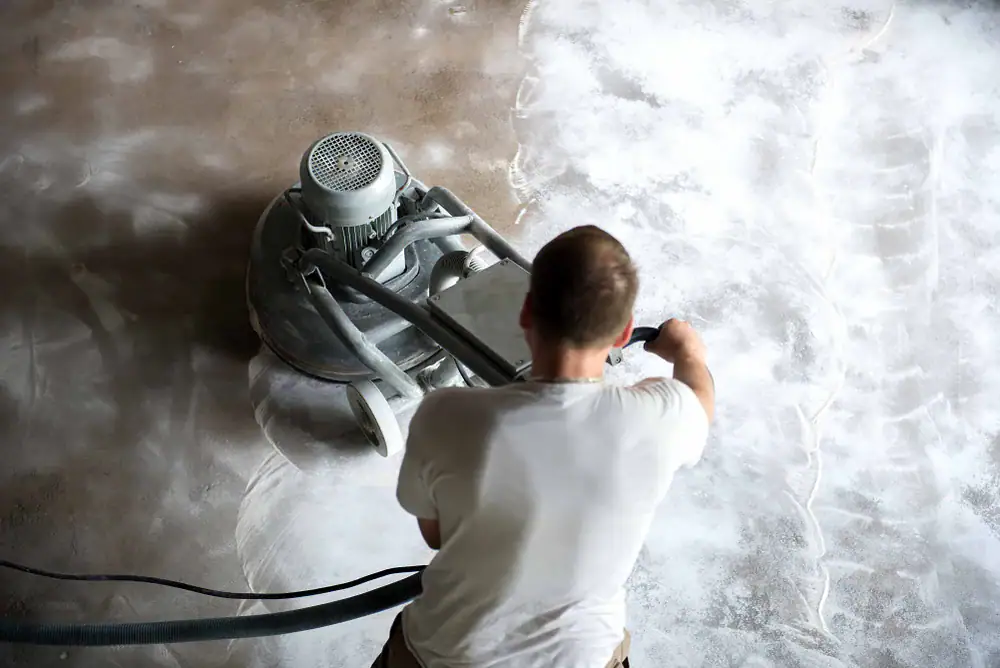 A person in a white shirt operates a floor grinding machine, smoothing or polishing a concrete surface covered in white dust or powder. The image is taken from above, showing the process in progress.
