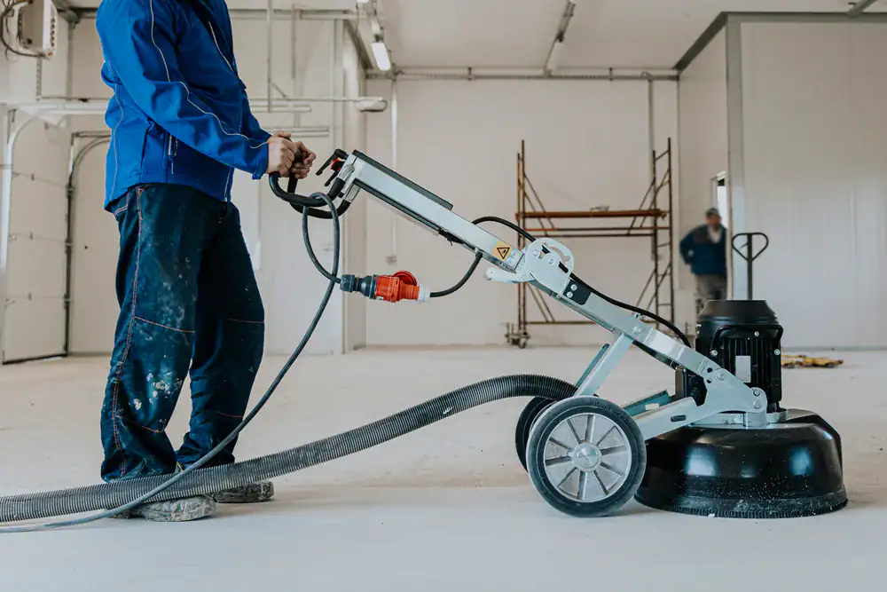 A worker in a blue jacket operates a floor grinding machine inside a large, unfinished room with white walls and construction materials visible in the background.
