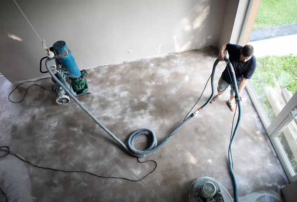 A person operating a floor sanding machine on a dusty, unfinished floor in a bright room with large windows and a view of grass outside.