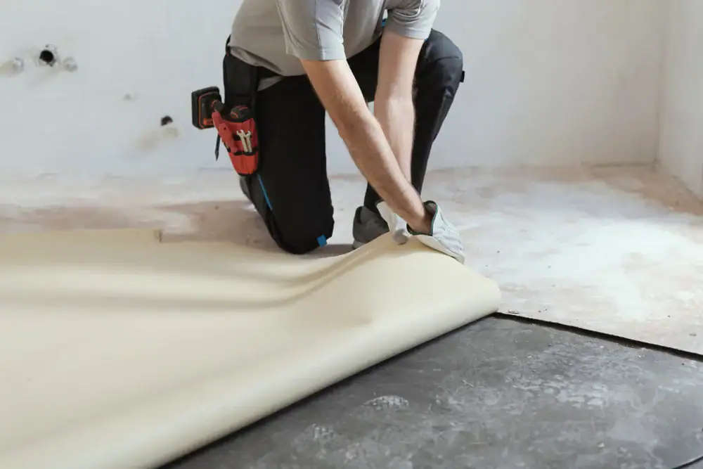 A person wearing gloves and work clothes is installing beige vinyl flooring over a black subfloor in a room under renovation. Tools are attached to their belt, and the walls are unfinished.