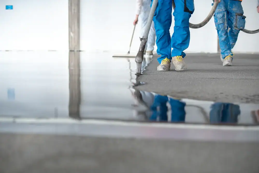 Workers in blue protective clothing are applying and smoothing wet concrete or liquid flooring on a large indoor surface. The floor is glossy and reflective, and equipment hoses are visible.