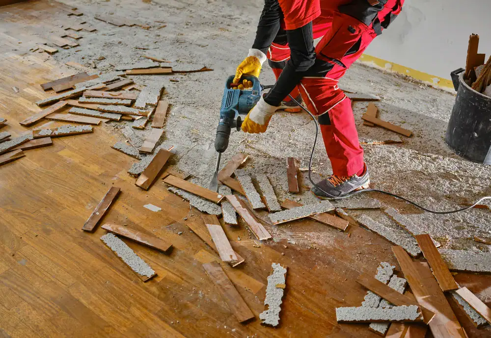 A person in red work clothes uses a power tool to remove damaged wooden planks and concrete debris from a floor during a renovation project.