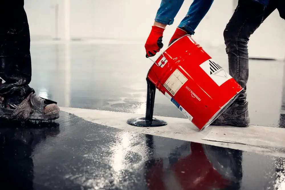 Two workers pour black liquid from a red metal container onto a shiny floor. One worker wears red gloves and blue sleeves; both wear work boots. The scene suggests flooring or construction work.