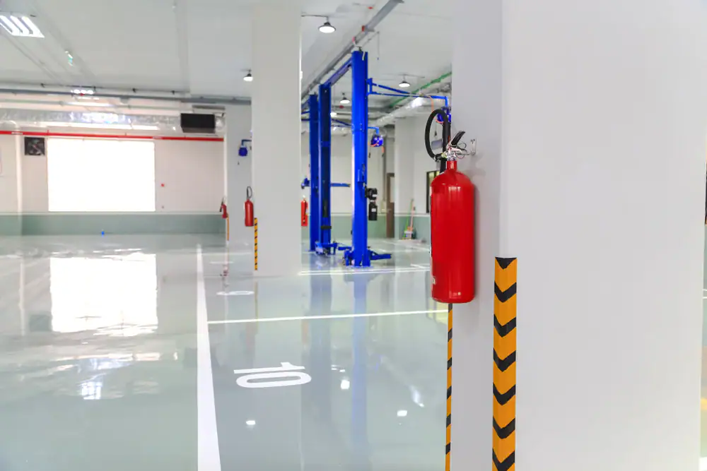A clean, modern indoor parking garage with shiny floors, numbered parking spaces, blue vehicle lifts, and red fire extinguishers mounted on white columns marked with yellow and black safety stripes.