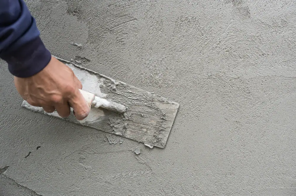 A close-up of a hand using a trowel to spread and smooth wet cement or plaster on a wall, creating an even surface.