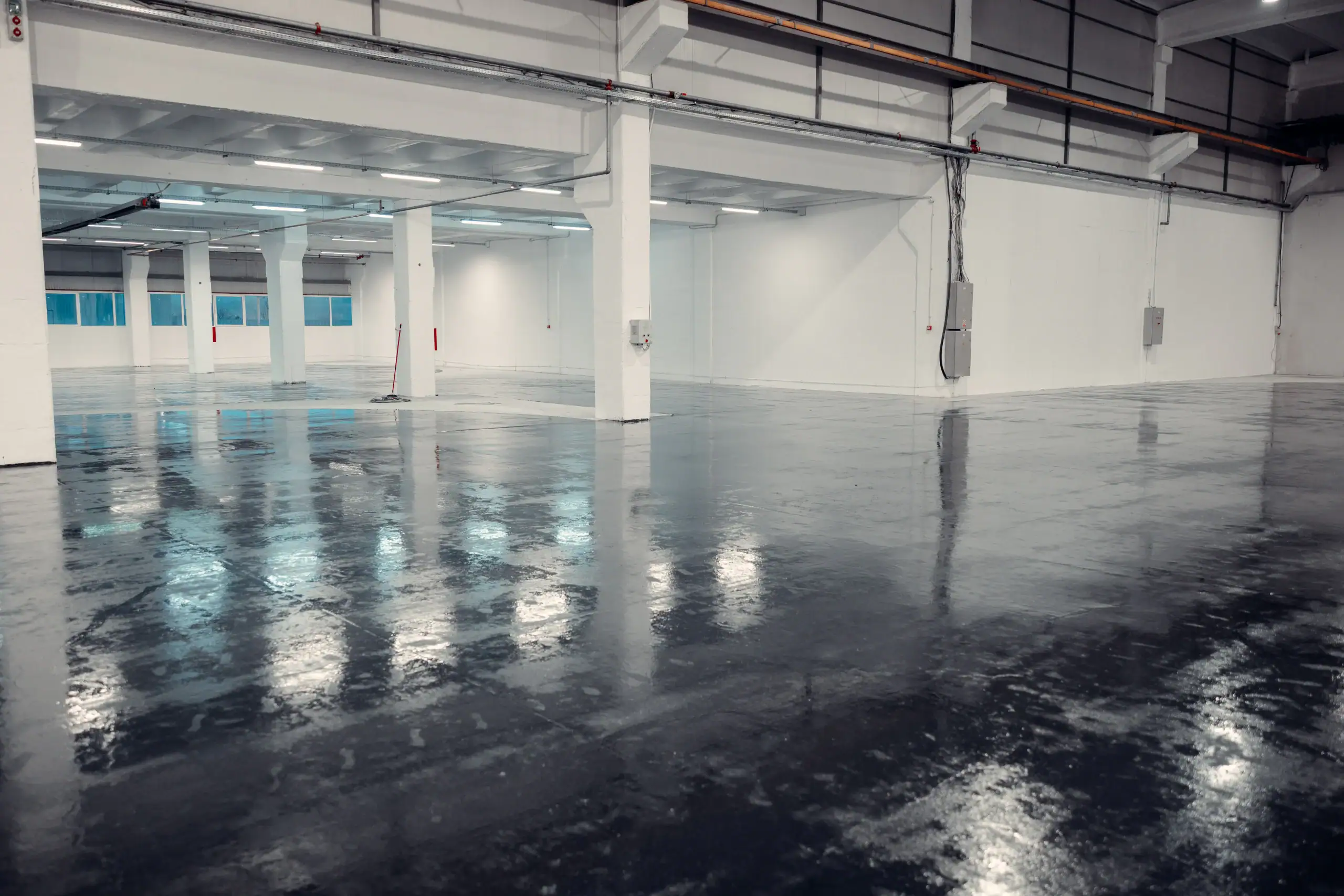Large, empty industrial warehouse with white walls, pillars, and a shiny, reflective concrete floor. The space is well-lit with overhead fluorescent lights.