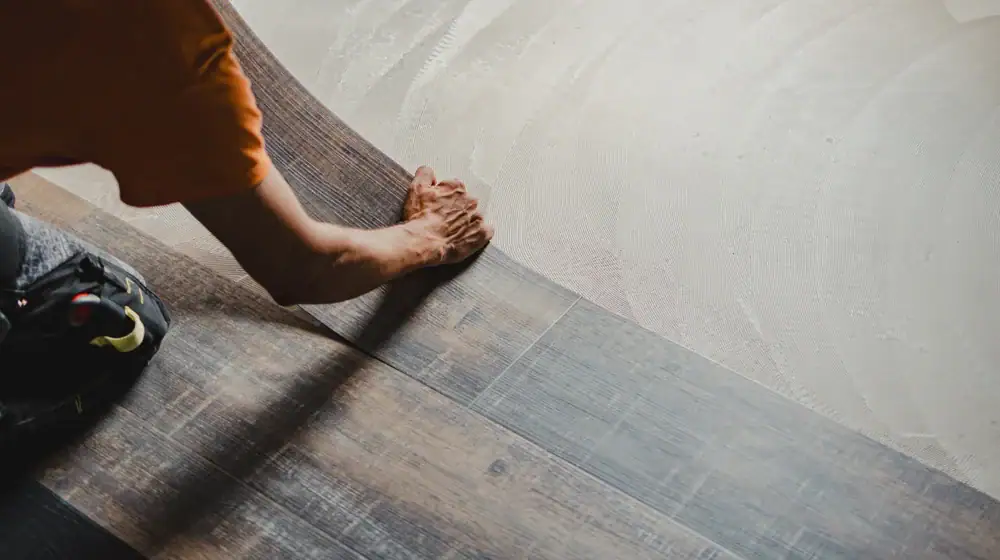 A person installs dark wood-look vinyl flooring, pressing the plank securely onto adhesive spread over the subfloor with one hand while wearing an orange shirt and kneeling on the floor.
