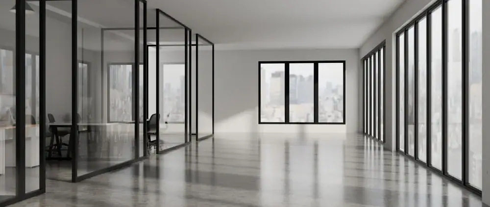 A modern, empty office space with large glass windows, glass-walled meeting rooms, and polished concrete floors. City buildings can be seen through the windows, and a few chairs and desks are visible in the meeting rooms.