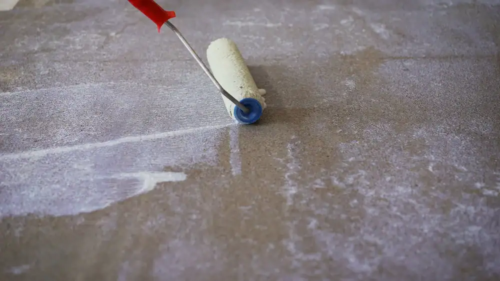 A paint roller with a red handle is being used to apply a light-colored coating to a rough, gray surface. The roller leaves visible streaks as it moves across the surface.