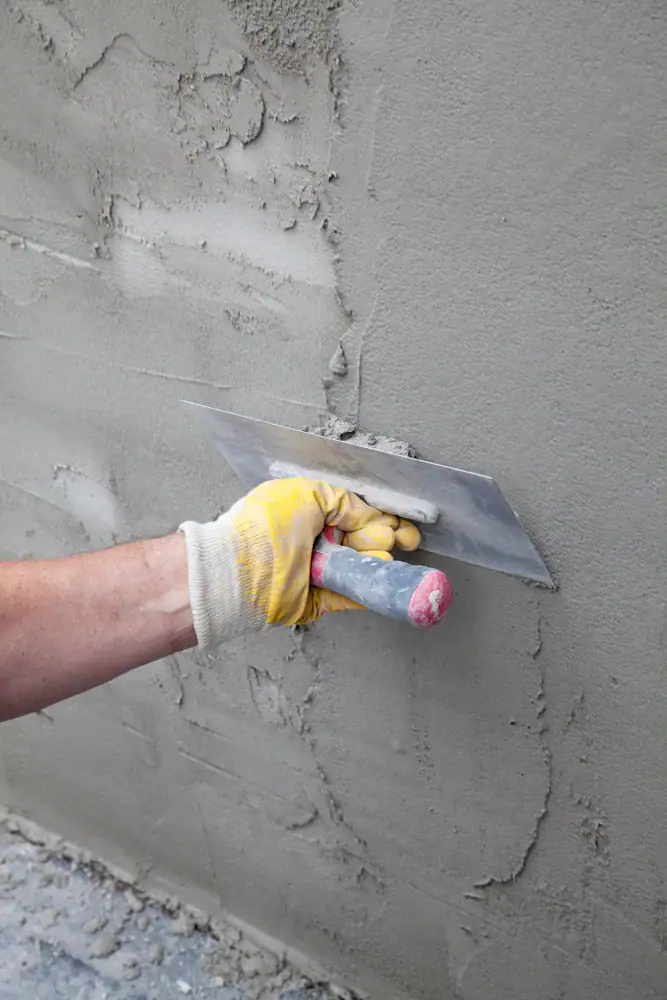 A person wearing a yellow work glove uses a trowel to smooth wet cement on a wall, creating an even surface.