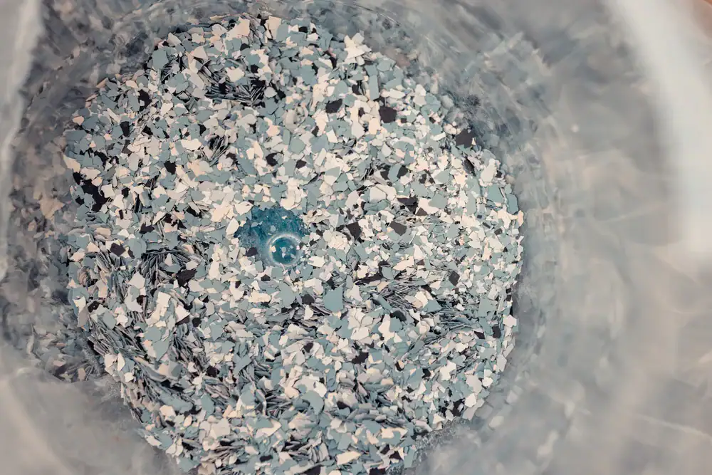 A close-up, top-down view of small, shredded pieces of blue and white plastic inside a transparent container, resembling recycled plastic flakes ready for processing.