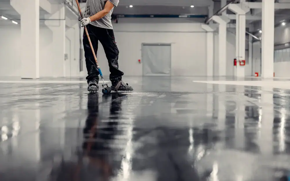 A person wearing gloves and work clothes uses a large roller to apply a shiny coating to the floor of a spacious, empty industrial building with white walls and columns.