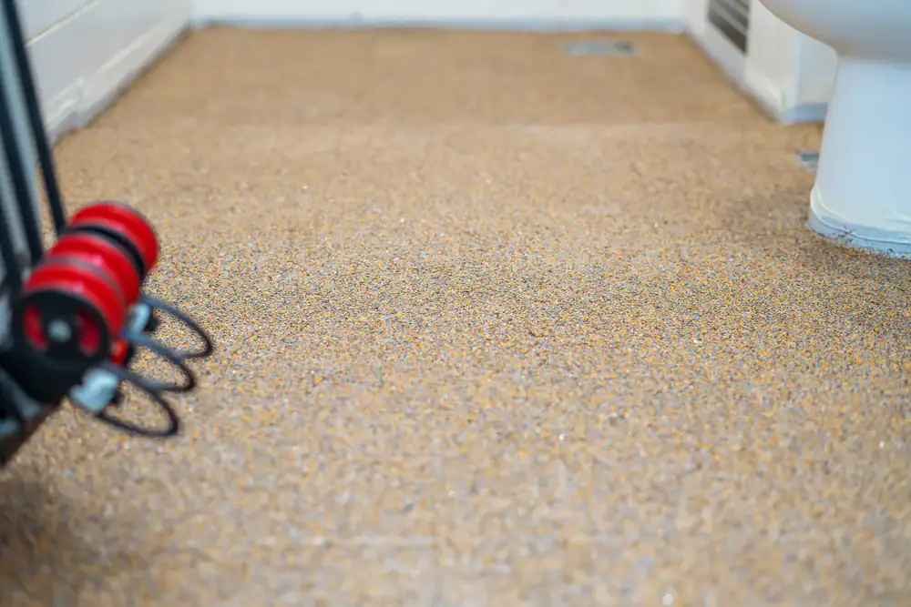 Close-up of a bathroom floor covered with a speckled, textured, tan-colored surface. A white toilet is partly visible on the right, and part of a black object with red wheels is on the left.