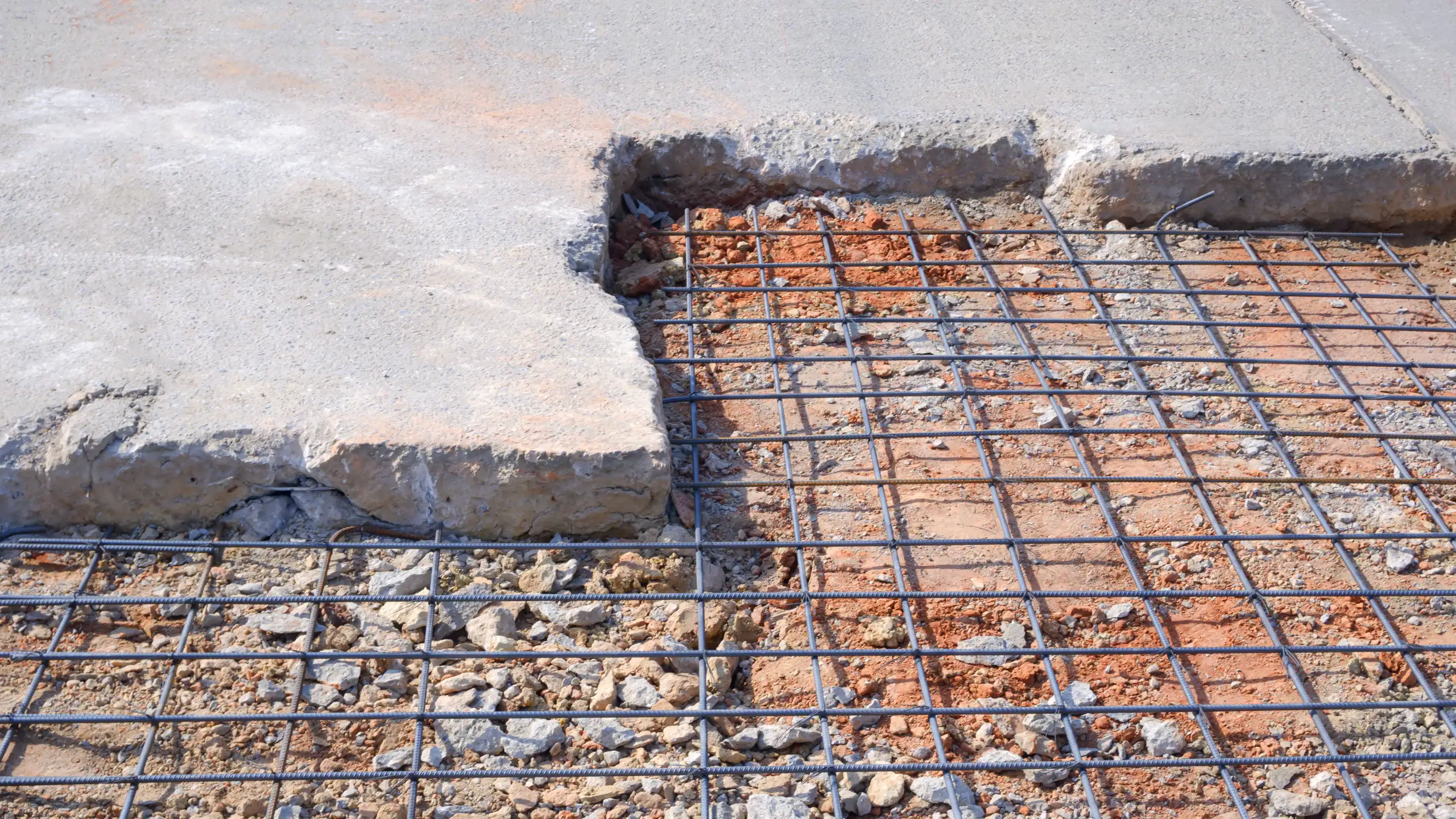 Concrete slab partially removed, revealing a grid of steel reinforcement bars and crushed rocks beneath the surface, exposing the construction layers of the pavement.