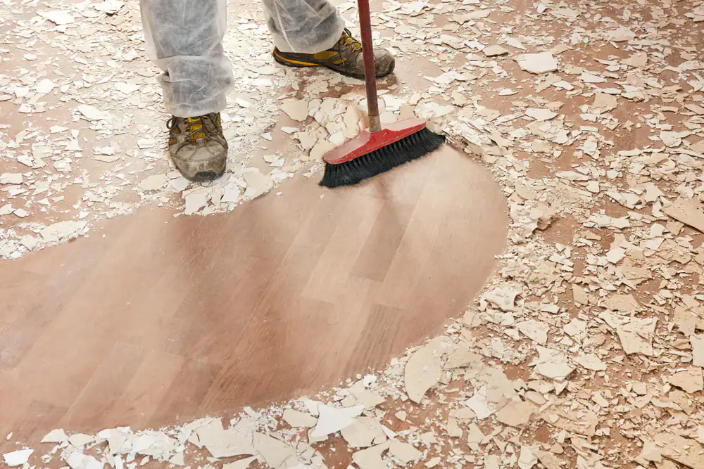 A person in work clothes and boots sweeps broken drywall debris from a wooden floor with a red broom, clearing a path through the dust and rubble.