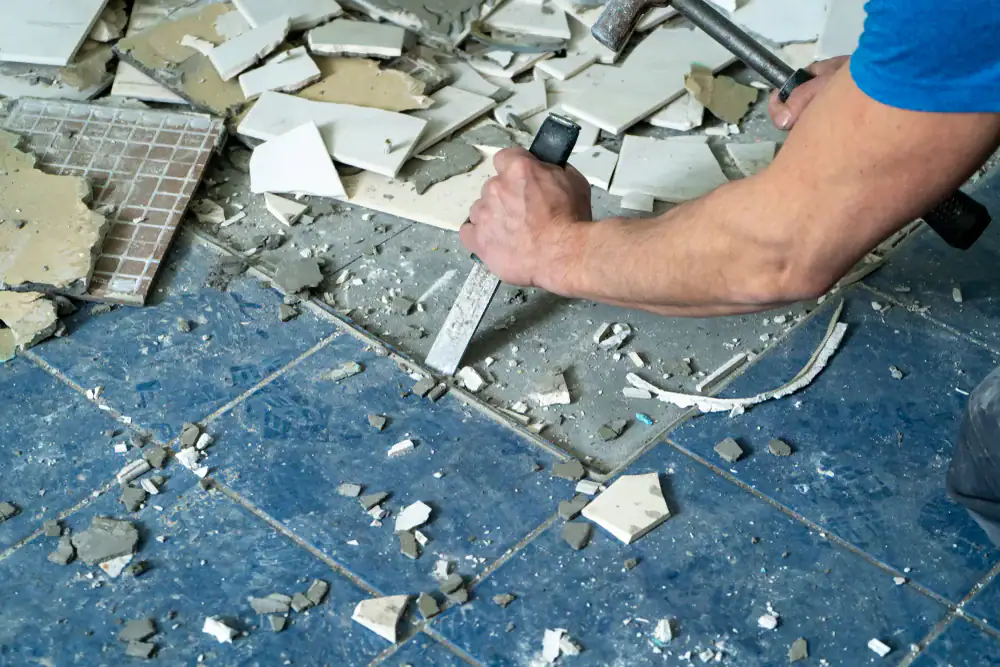 A person uses a chisel and hammer to remove broken white tiles from a floor, with blue tiles and scattered debris visible.