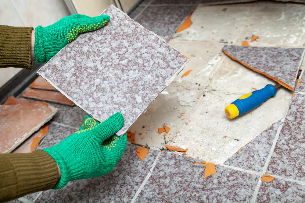 A person wearing green gloves is removing a floor tile, holding a loosened tile above broken tiles and debris on the floor; a tool with a blue and yellow handle lies nearby.