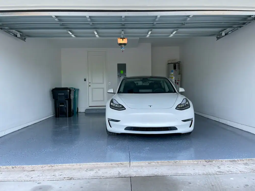 A white Tesla Model 3 is parked inside a clean, spacious two-car garage with a gray floor, closed trash bins on the left, and a refrigerator on the right.