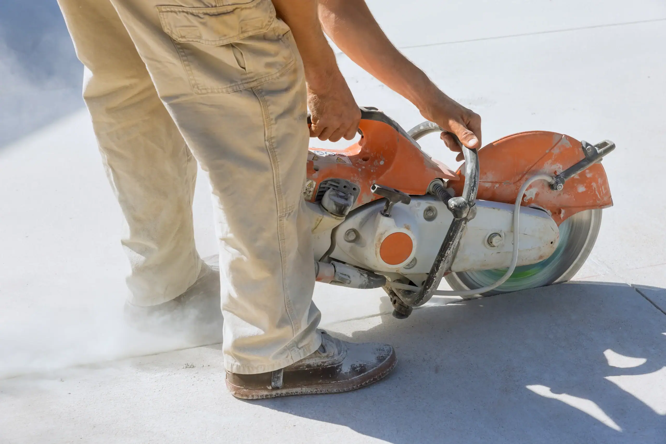 A person wearing beige pants and work shoes cuts concrete with an orange circular saw, creating dust on a bright outdoor surface.