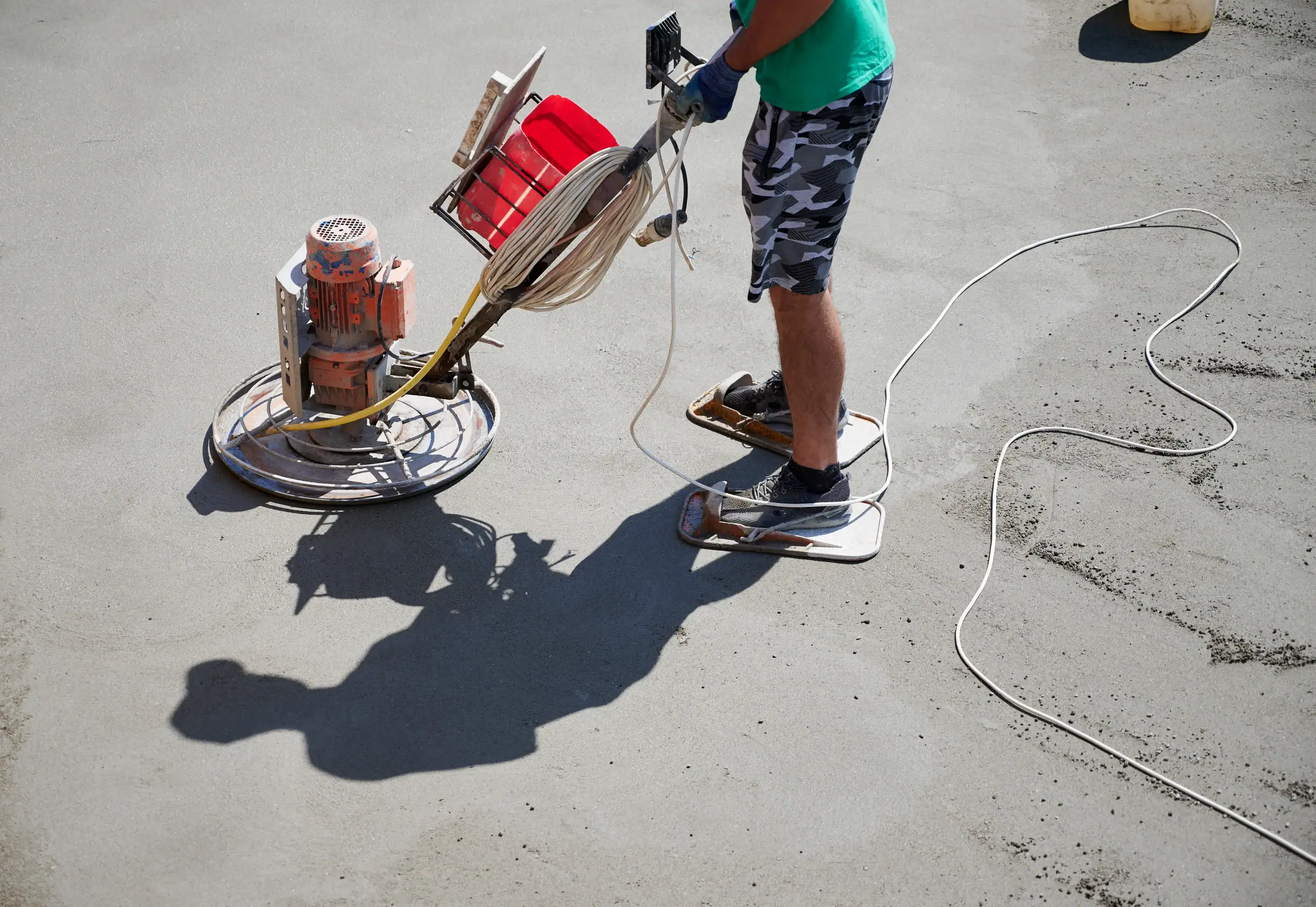 A worker in camouflage shorts operates a power trowel on a concrete surface, wearing makeshift boards on his feet to avoid leaving footprints. His shadow is cast on the smooth concrete.