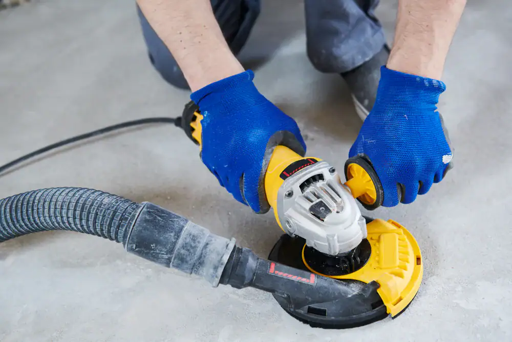 A person wearing blue gloves uses a yellow and black power tool connected to a vacuum hose to sand or grind a concrete floor.