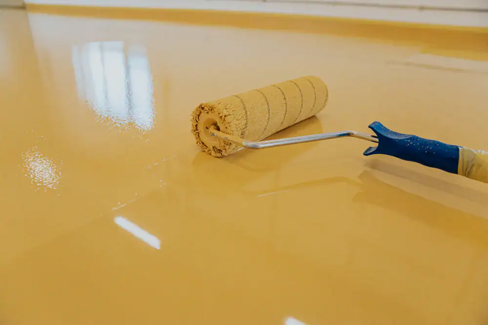 A paint roller is being used to apply a smooth, glossy yellow coating to a floor, creating a shiny and even surface. Reflections of windows are visible on the wet paint.